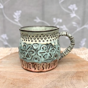 May include: A ceramic mug featuring a light blue and cream glaze with embossed floral patterns and dotted textures. The mug has a rounded body, a dark rim, and a textured handle. The base of the mug transitions into a warm, earthy peach tone.