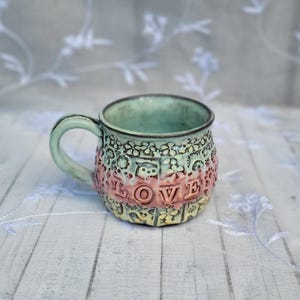 May include: A ceramic mug with a light green handle and interior. The exterior features embossed floral patterns and the word "LOVE" in pink letters. The mug has a textured, handcrafted appearance.