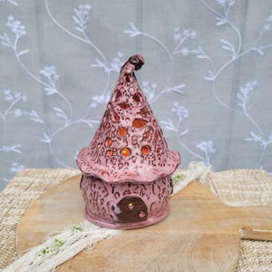 May include: A pink ceramic fairy house with a textured surface and a pointed roof. The roof has cut-out holes that allow light to shine through. The house features a small, brown door and is designed to hold a candle or small light.
