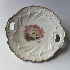 May include: White porcelain serving dish with a gold rim and handles. The center features a floral and bird design in pink, green, and black. The dish has a scalloped edge and a textured surface.