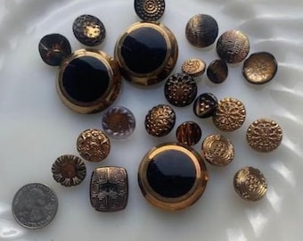 Puede incluir: Una colección de botones antiguos de varios tamaños y diseños. Los botones presentan una mezcla de colores dorados, marrones y negros. Algunos botones tienen intrincados patrones en relieve, mientras que otros son lisos o tienen un acabado brillante. Se incluye una moneda para referencia de tamaño.