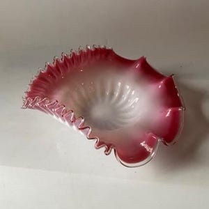 May include: A ruffled-edge glass bowl with a white interior and a gradient of pink to red on the exterior. The bowl has a unique, wavy shape and a delicate, textured rim.