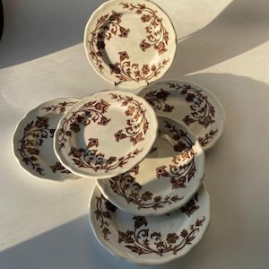 May include: A set of five white ceramic plates with scalloped edges, each decorated with a brown floral pattern featuring swirling vines and blossoms. The plates are arranged in a stacked and overlapping formation, showcasing the consistent design.