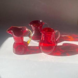 May include: Three ruby red glass pitchers with handles are arranged on a white surface. The pitcher on the left has a yellow handle. The pitchers are of varying sizes and shapes, with a glossy finish.