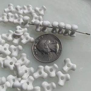 Puede incluir: Primer plano de un montón de cuentas de cerámica blanca con un diseño de tres puntas. Las cuentas están esparcidas sobre una superficie blanca junto a una moneda de 25 centavos de dólar estadounidense.