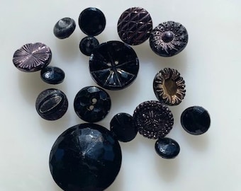 May include: A collection of vintage black and purple buttons, some with intricate designs, on a white surface.