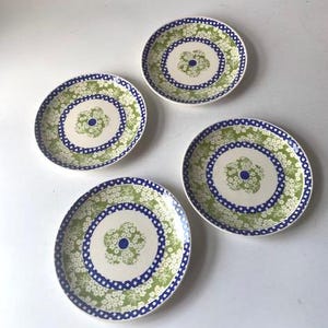 May include: Four round ceramic plates featuring a central flower motif surrounded by a blue and white checkered border. The outer rim displays a dense pattern of small white flowers on a light green background, creating a consistent design across all pieces.