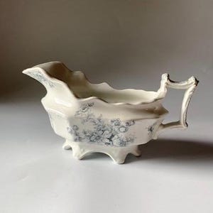 May include: A white porcelain gravy boat with a blue floral pattern and a handle. The boat has a scalloped edge and three feet.