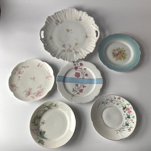 May include: A collection of six antique porcelain plates with floral designs. The plates vary in size and shape, featuring scalloped edges and delicate patterns in pink, blue, and green. One plate has a blue stripe, and another has handles.