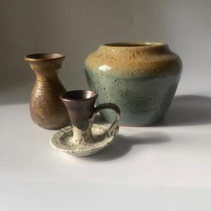May include: A still life of three ceramic pieces. A small brown vase, a candle holder with a handle, and a large vase with a blue-green base and a tan top. The candle holder is white with brown speckles.
