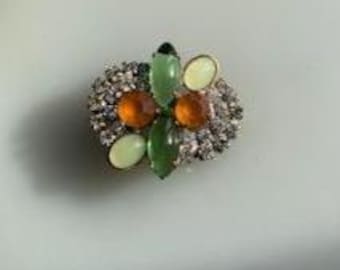May include: A vintage brooch with a cluster of colorful gemstones. The brooch features a central arrangement of green and orange stones, surrounded by smaller clear crystals. The design is set in a silver-toned metal.