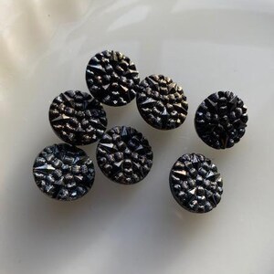 Puede incluir: Un conjunto de siete botones negros texturizados con un diseño facetado. Los botones redondos tienen un brillo ligeramente iridiscente, dispersos sobre una superficie blanca.