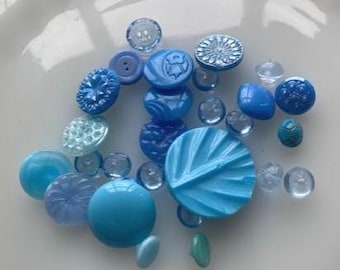 May include: A collection of blue buttons in various shades and sizes, including round, oval, and faceted designs. Some buttons are clear, while others are solid or have embossed patterns. The buttons are scattered on a white surface.