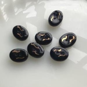Könnte beinhalten: Sieben glänzende, ovale schwarze Knöpfe mit einem goldfarbenen, geschwungenen Design. Jeder Knopf hat zwei kleine Löcher zum Annähen. Diese dekorativen Knöpfe eignen sich für verschiedene Nähprojekte. Die Knöpfe sind ca. 1,5 cm lang.