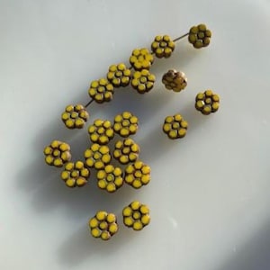 Könnte beinhalten: Eine Sammlung gelb-brauner, blütenförmiger Perlen. Einige Perlen sind auf einem Draht aufgereiht, andere verstreut. Die Perlen haben ein detailliertes, strukturiertes Aussehen mit gelben Zentren und braunen Blütenblättern. Geeignet für die Schmuckherstellung.