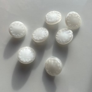 Puede incluir: Un conjunto de siete botones blancos ovalados con borde festoneado. Los botones tienen un brillo nacarado y están dispersos sobre una superficie clara. Ideales para costura y manualidades, añadiendo un toque decorativo.