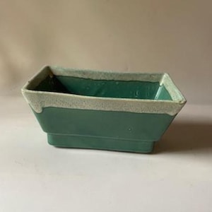May include: A rectangular, green ceramic planter with a light-colored, textured rim. The planter has a stepped base and a slightly flared shape. The interior is a darker shade of green.