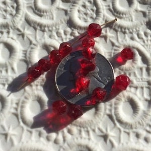 May include: A group of small, red, faceted glass beads, some on a gold pin, next to a US quarter for scale.