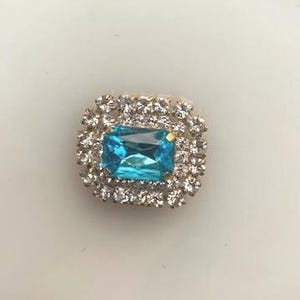 Op de afbeelding: Een vierkante, sierlijke strass-knop met een grote, rechthoekige, lichtblauwe middensteen. De middensteen is omgeven door een halo van kleinere, heldere strass-steentjes, gezet in een goudkleurige metalen basis. Waarschijnlijk voor decoratieve doeleinden.