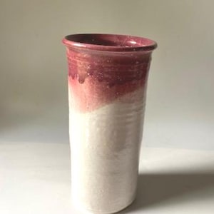 May include: A handmade ceramic vase with a cylindrical shape. The top half is a deep burgundy color, transitioning to a creamy white at the base. The vase has a slightly flared rim and a textured surface, showcasing the craftsmanship.