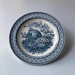 May include: A blue and white decorative plate with a floral and architectural design. The plate features a detailed illustration of flowers and a building, surrounded by a patterned border. The plate is likely made of ceramic.