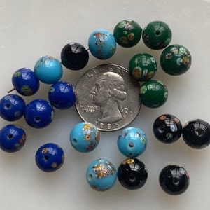 Op de afbeelding: Een verzameling vintage glazen kralen in verschillende kleuren, waaronder blauw, groen en zwart. Sommige kralen hebben bloemmotieven, terwijl andere effen kleuren hebben. De kralen zijn gerangschikt op een wit oppervlak.