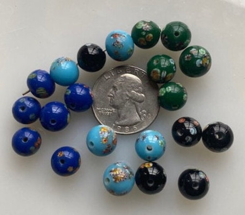 Op de afbeelding: Een verzameling vintage glazen kralen in verschillende kleuren, waaronder blauw, groen en zwart. Sommige kralen hebben bloemmotieven, terwijl andere effen kleuren hebben. De kralen zijn gerangschikt op een wit oppervlak.