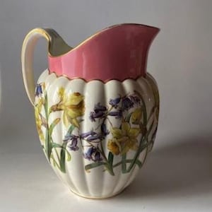 May include: A decorative ceramic pitcher with a cream-colored base and a pink rim. The pitcher is adorned with a floral pattern featuring yellow and purple flowers. The handle and rim have gold accents.