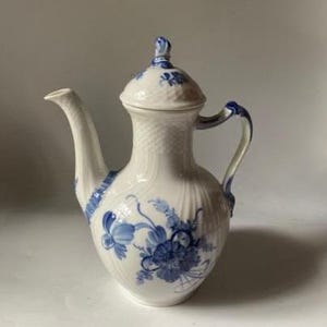 May include: A white porcelain teapot featuring a delicate blue floral pattern painted on the body. The teapot has a curved spout, a handle with blue accents, and a matching lid with a textured basket-weave design on the neck and lid.