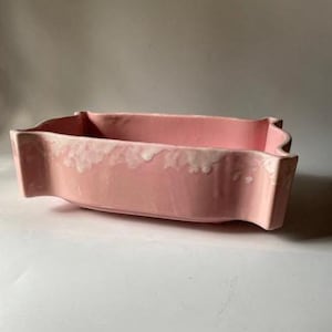 May include: A rectangular, pink ceramic planter with a scalloped edge and a white, textured trim. The planter has a curved design and is suitable for indoor or outdoor use. The overall aesthetic is vintage and decorative.