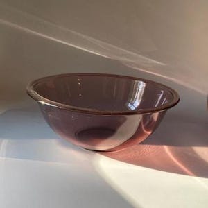 May include: A clear, lavender-colored glass mixing bowl. The bowl has a rounded shape with a slightly flared rim. The light catches the glass, creating highlights and shadows.