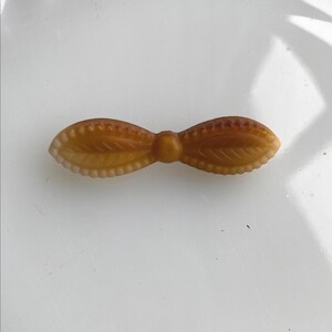 May include: A vintage-style, amber-colored bow-shaped brooch. The brooch features a detailed leaf design on each side of the bow, with a textured edge. The bow is made of a translucent material, and the overall design is symmetrical.