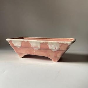 May include: A rectangular, pink ceramic planter with a white rim and three small feet. The planter has a ridged design on the sides and is suitable for indoor or outdoor use. The planter is approximately 12 inches long.