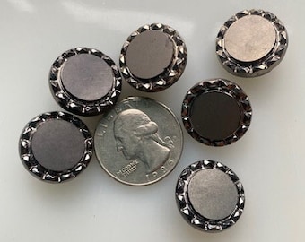 Op de afbeelding: Zes ronde zwarte en zilveren knopen met een verhoogde rand en een glad, plat midden. De knopen zijn in een halve cirkel op een wit oppervlak gerangschikt.