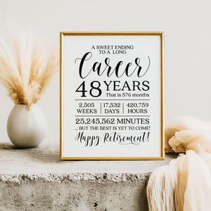 May include: A framed print with a gold border, featuring the words "Career 48 Years" and "Happy Retirement!" in elegant script. The print includes the equivalent of months, weeks, days, hours, and minutes. A vase with dried flowers is to the left.