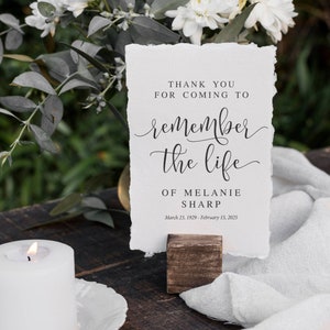 May include: A white card with black text on a wooden table with white flowers and a white candle. The text reads "Thank you for coming to remember the life of Melanie Sharp, March 23, 1929 - February 15, 2025".