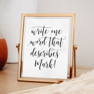 May include: A gold-framed sign with a white background and black calligraphy that reads "write one word that describes Mark!"