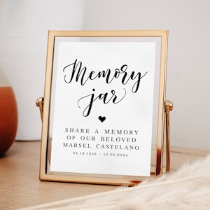 May include: A gold-framed sign with the text "Memory Jar" in black cursive font. Below the text is a small black heart and the words "Share a memory of our beloved Marsel Castelono 05.19.1956 - 12.05.2024".