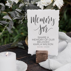 May include: A white card with black text that reads "Memory Jar" and "Share a memory of our beloved Karla Wilson 08.05.1954 - 10.06.2025". The card is propped up on a small wooden stand. A white candle is in the foreground.