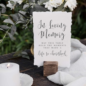 May include: A white card with the text "In Loving Memory" in elegant script, held by a small wooden block. A lit white candle and white flowers are in the background, creating a serene atmosphere.