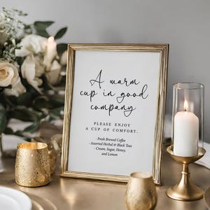 May include: A framed sign with the text "A warm cup in good company" sits on a table, next to a candle in a gold holder and a bouquet of white roses. The sign also lists coffee and tea options.