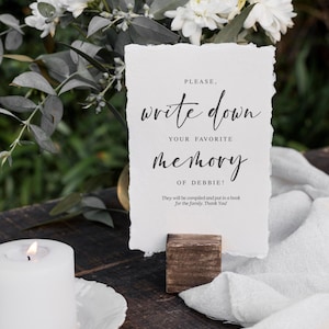 May include: A white card with black text on a wooden stand. The text reads "Please, write down your favorite memory of Debbie! They will be compiled and put in a book for the family. Thank You!"