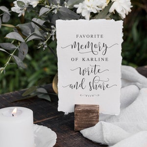 May include: A white card with black text on a wooden stand. The text reads "Favorite Memory of Karline write and share". The card is surrounded by white flowers and a white cloth.