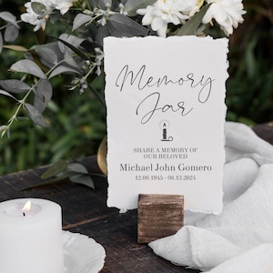 May include: A white card with black text that reads "Memory Jar" with a small candle icon. The text below reads "Share a memory of our beloved Michael John Gomero 12.09.1945 - 08.13.2024". The card is on a wooden stand.