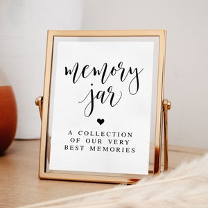 May include: A gold-framed sign with the text "memory jar" in black calligraphy. Below the text is a small black heart and the words "A collection of our very best memories".