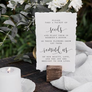 May include: A white card with black text on a wooden stand. The text reads "Please take a packet of seeds and plant them in Debbie's honour. As these flowers grow let them remind us of her beauty, love, and the joy she brought to our lives."