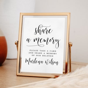 May include: A gold-framed sign with a white background and black text that reads "share a memory" and "Please take a card and share a memory of our beloved Marlena Wilson".