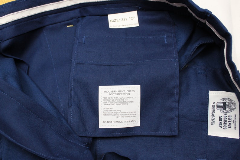 Puede incluir: Un pantal&oacute;n de vestir azul marino con una etiqueta blanca que dice "Trousers, Men's, Dress, Polyesterwool" y una etiqueta blanca con el texto "Defense Logistics Agency by GSA Apparel Incorporated".