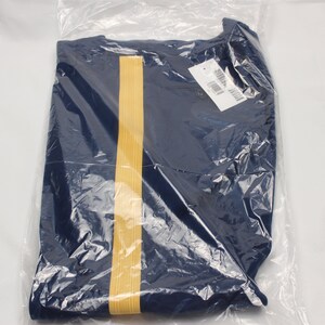 Puede incluir: Una camisa azul marino con una franja dorada en la parte delantera. La camisa est&aacute; doblada y empaquetada en una bolsa de pl&aacute;stico transparente.