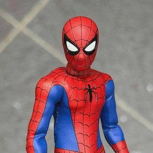 Puede incluir: Una figura de acción de Spider-Man con un traje de tela rojo y azul que presenta un patrón de telaraña negro impreso en las secciones rojas. La figura tiene un emblema de araña negro en el pecho, lentes oculares blancos con bordes negros y articulaciones visibles.